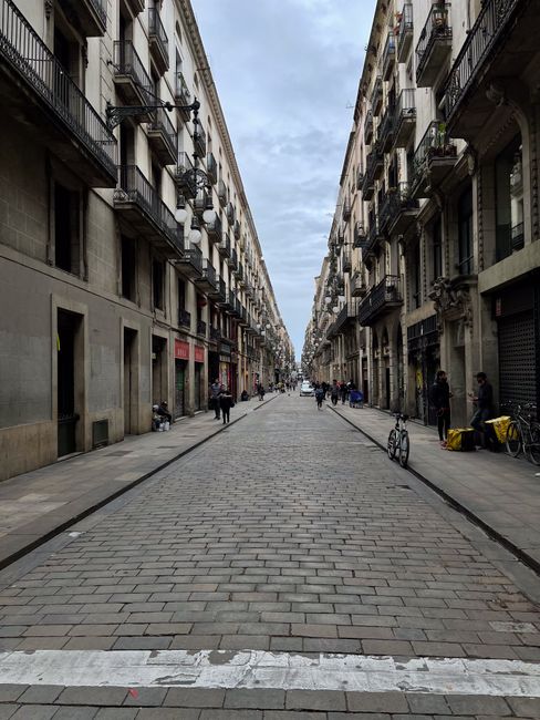A wide street view in Eixample Barcelona showing a historic multi-storey building with ornate architectural details, balconies, and a domed rooftop. Parked vehicles, including a white delivery van wit