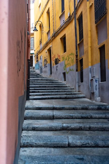 A narrow outdoor staircase in a residential area with concrete steps ascending between closely spaced multi-storey buildings. On the left, windows with decorative metal bars are visible, alongside sma